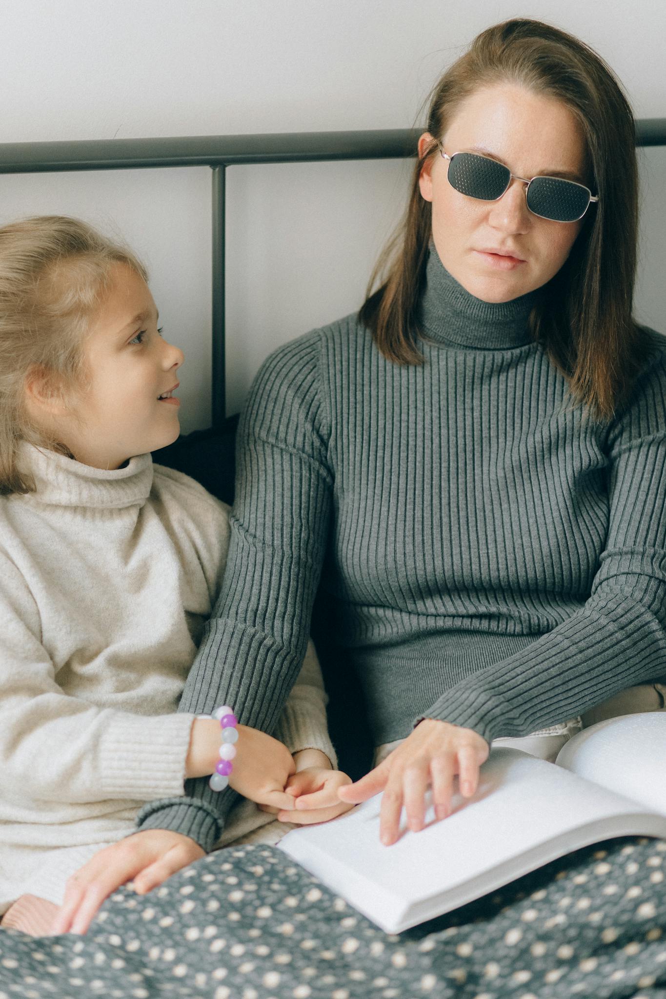 Woman in sunglasses reading braille with a girl on bed, depicting learning with disability.